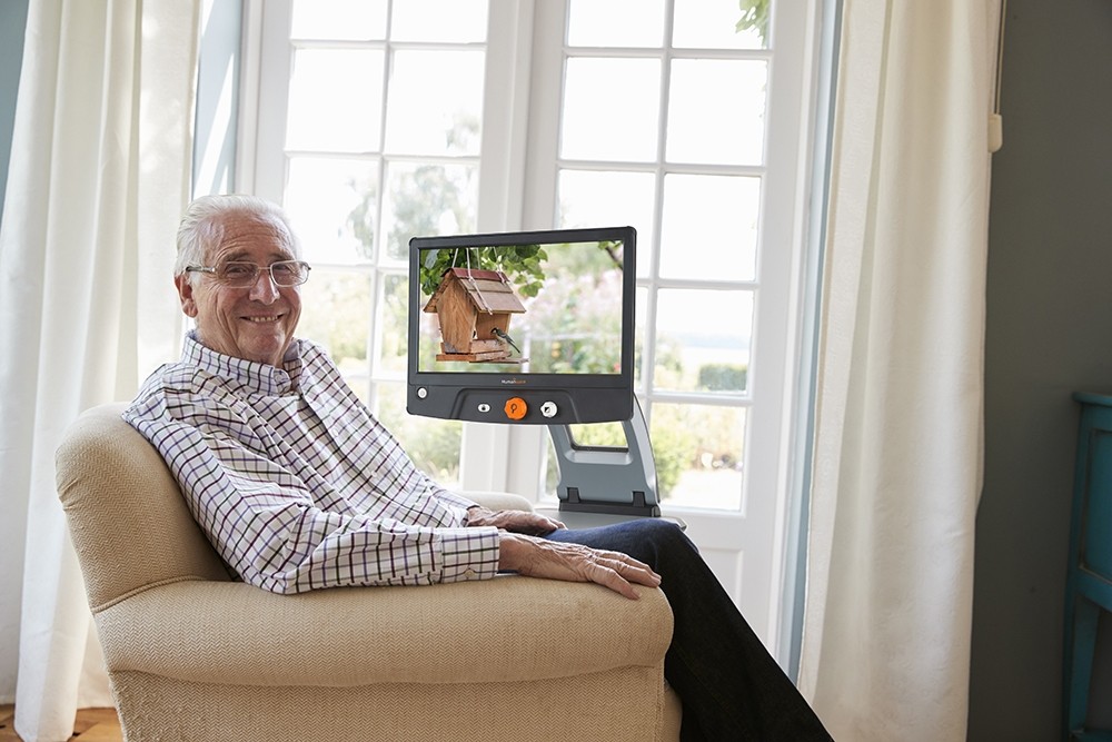 man sitting in chair smiling looking at camera. Reveal 16 digital magnifier next to chair with crystal clear photo of bird house and tree.